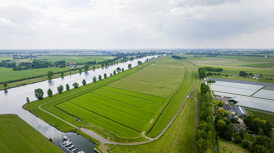 Luchtaanzicht van de Genderensche uiterwaard die uit een groot stuk grasland bestaat en rechts grenst aan een boerderij en links aan de Bergsche Maas. Aan de stroomopwaartse ligt een kleine jachthaven.
